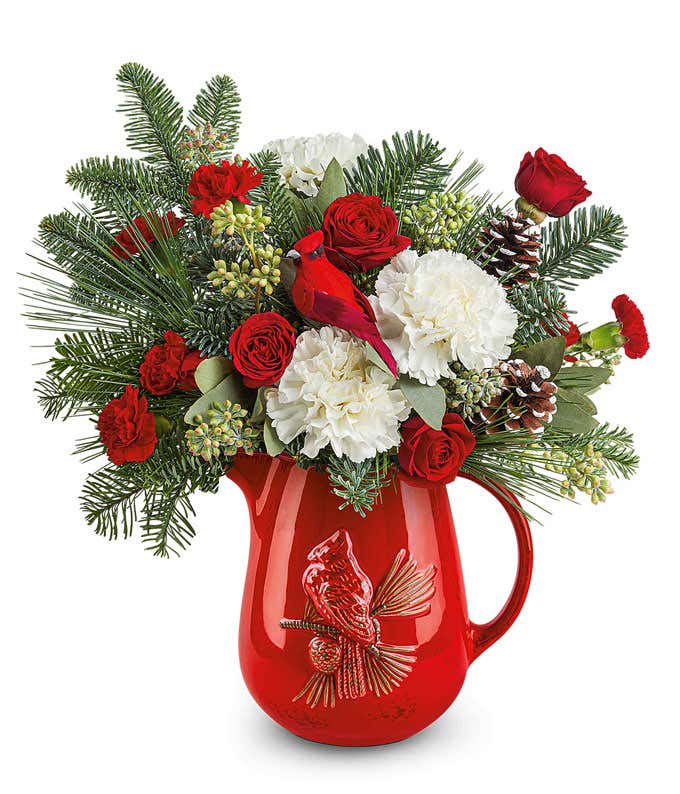 Holiday floral arrangement with white roses, red spray roses, red miniature carnations, pinecones, and greenery in a red ceramic pitcher featuring a raised cardinal design.