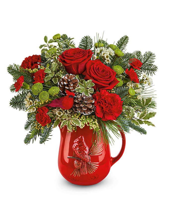 Christmas floral arrangement with red roses, red carnations, green button spray chrysanthemums, pinecones, greenery, and white waxflower in a red ceramic pitcher featuring a raised cardinal design.