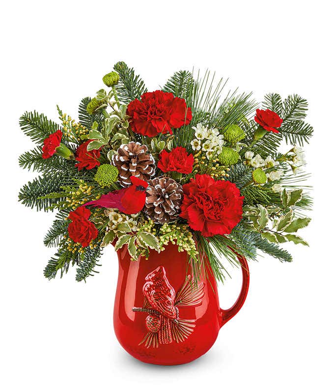 Christmas floral arrangement with red roses, red carnations, green button spray chrysanthemums, pinecones, greenery, and white waxflower in a red ceramic pitcher featuring a raised cardinal design.