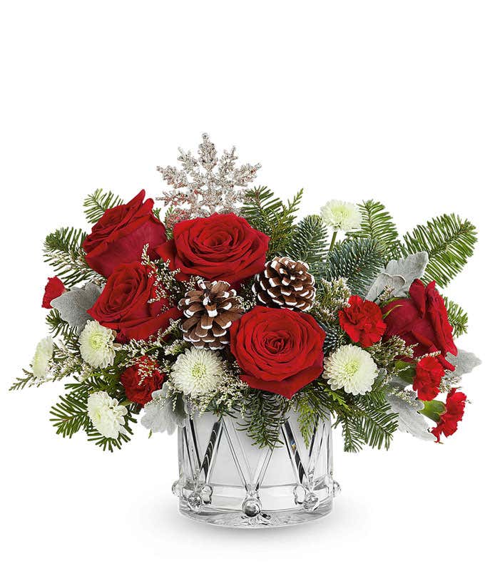 Holiday flower arrangement with red roses, miniature red carnations, and white button spray chrysanthemums in a silver drum-shaped container with pinecones, evergreens, and a glittery snowflake pick.