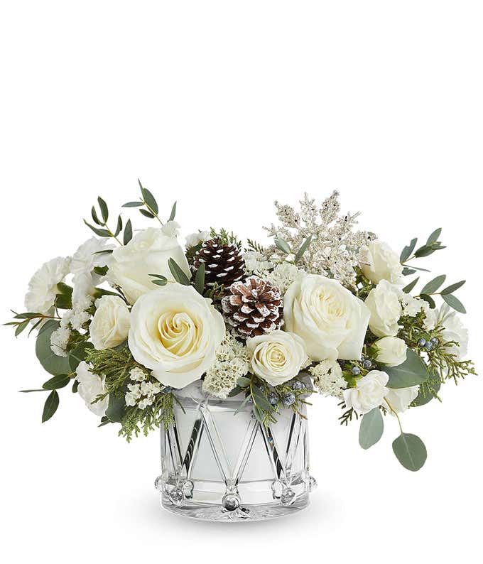Winter flower arrangement with white roses, spray roses, carnations, and mini carnations in a silver drum-shaped container with pinecones, greenery, and a glittery snowflake pick.