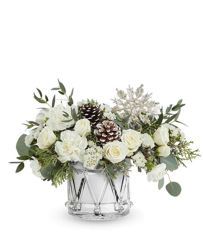 Winter flower arrangement with white roses, spray roses, carnations, and mini carnations in a silver drum-shaped container with pinecones, greenery, and a glittery snowflake pick.