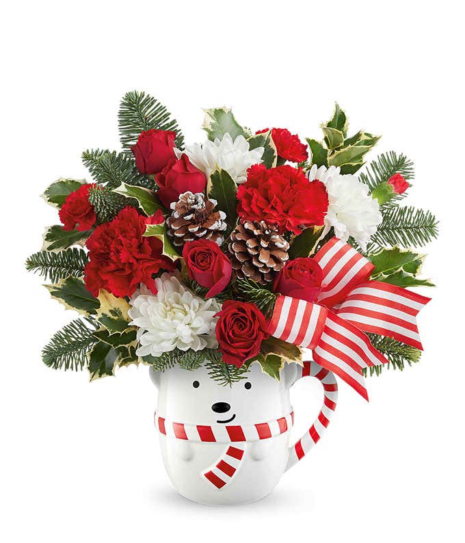 Festive holiday arrangement with red and white roses, red carnations, white mums, pinecones, greenery, and a red-and-white striped bow in a polar bear mug container.