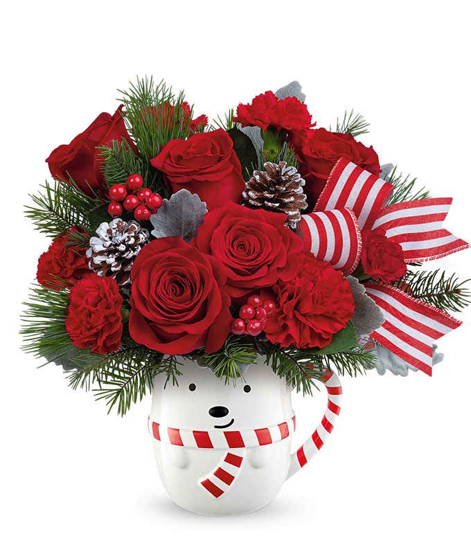 Holiday flower arrangement with red roses, red carnations, and mini red carnations in a white polar bear mug with pinecones, greenery, red berries, and a striped ribbon.