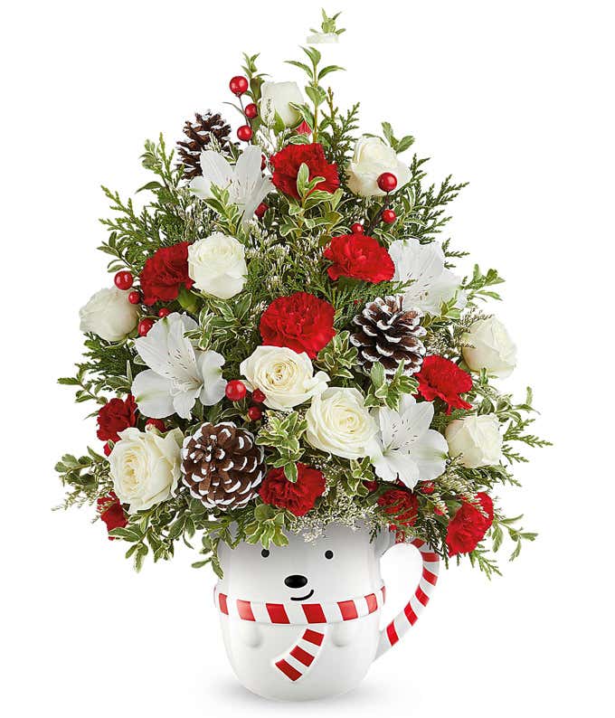 Christmas tree-shaped floral arrangement with red and white roses, red carnations, white alstroemeria, frosted pinecones, red berry accents, and greenery in a white bear mug with a red striped scarf.