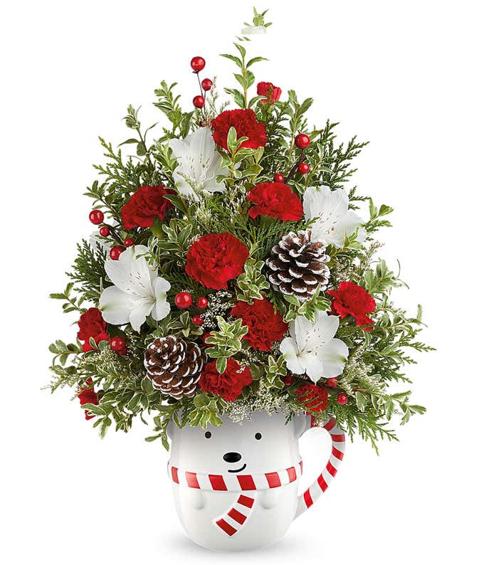 Christmas tree-shaped floral arrangement with red and white roses, red carnations, white alstroemeria, frosted pinecones, red berry accents, and greenery in a white bear mug with a red striped scarf.