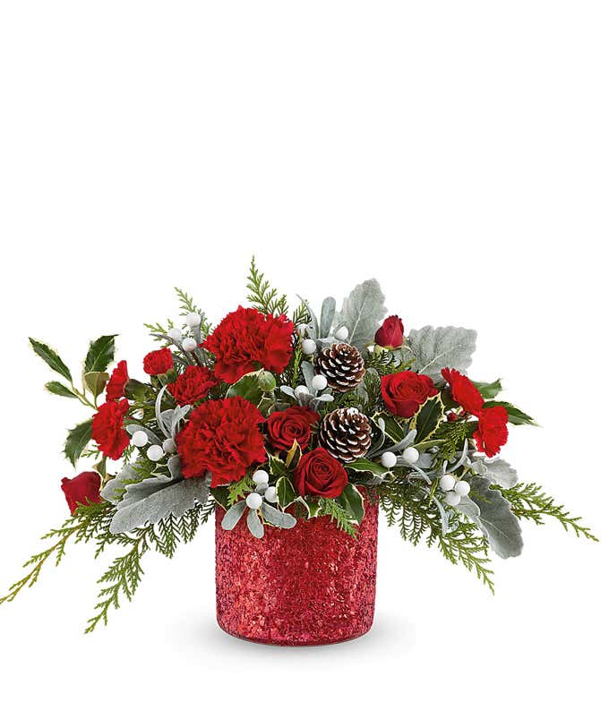 Holiday floral arrangement with red roses, red carnations, white berry picks, frosted pinecones, and seasonal greenery in a red glittered container.