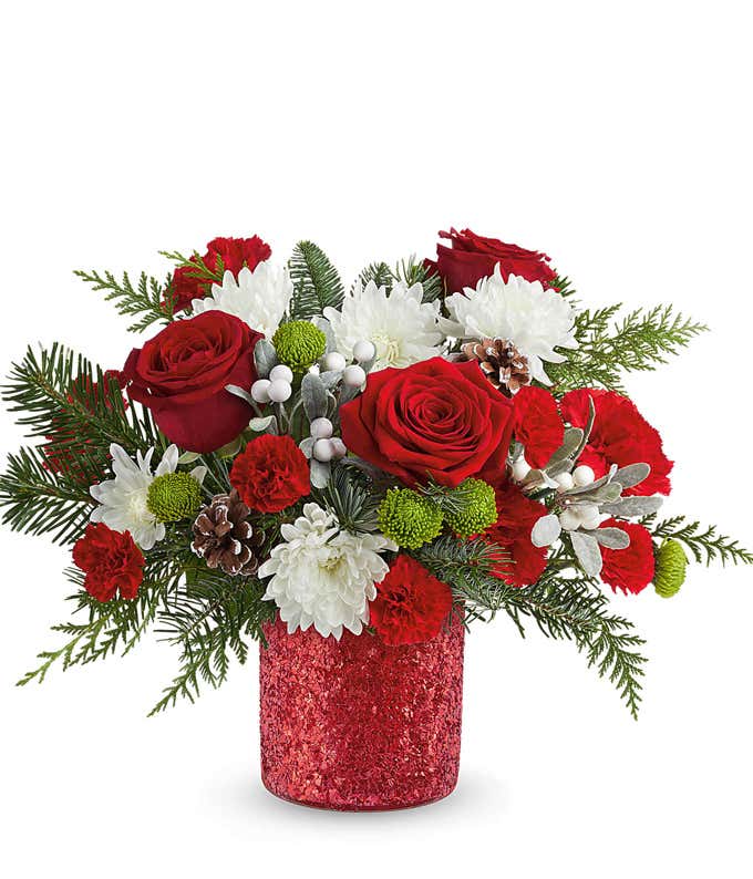 Holiday floral arrangement with red roses, red carnations, white cushion spray chrysanthemums, green button spray chrysanthemums, white berry picks, frosted pinecones, and seasonal greenery in a red glittered container.