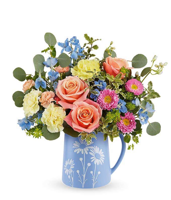 Floral arrangement of Peach Roses, Yellow carnations, peach mini carnations, light blue delphinium, and floral greenery in a pale blue pitcher vase with white daisy details