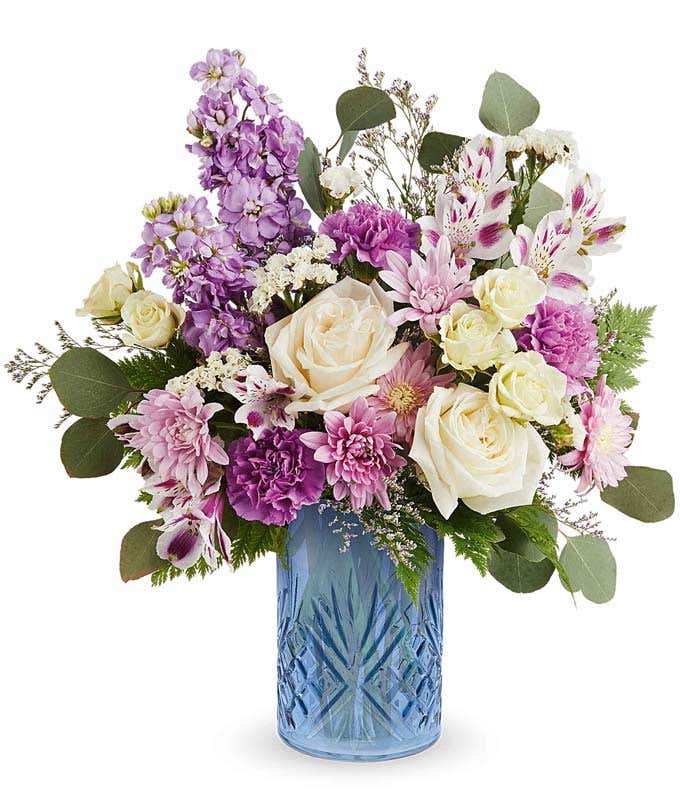 White roses, White Spray Roses, Lavender Alstroemeria, Purple Carnations, Lavender Stock, Lavender Cushion Stock, and Floral Greenery in a blue glass cylinder vase with design