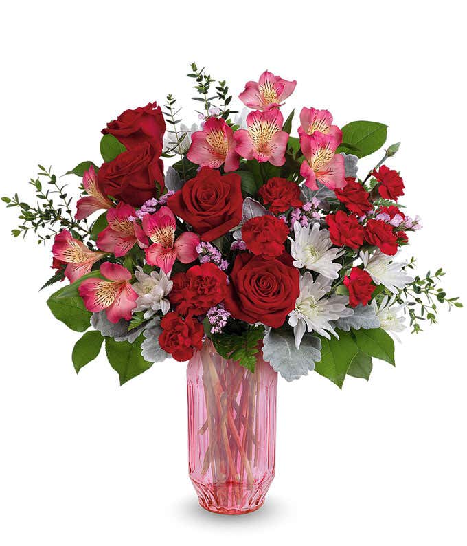 Valentine’s Day floral arrangement featuring red roses, red carnations, pink alstroemeria, white cushion mums, and lush greenery arranged in a pink glass vase.