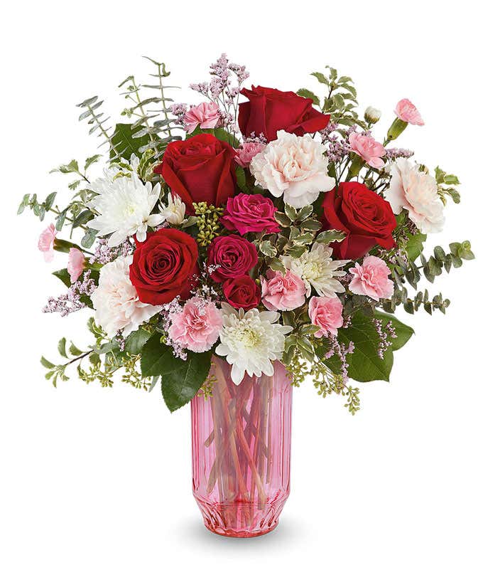 Valentine’s Day floral arrangement featuring red roses, blush and pink blooms, white flowers, and lush greenery arranged in a pink glass vase.
