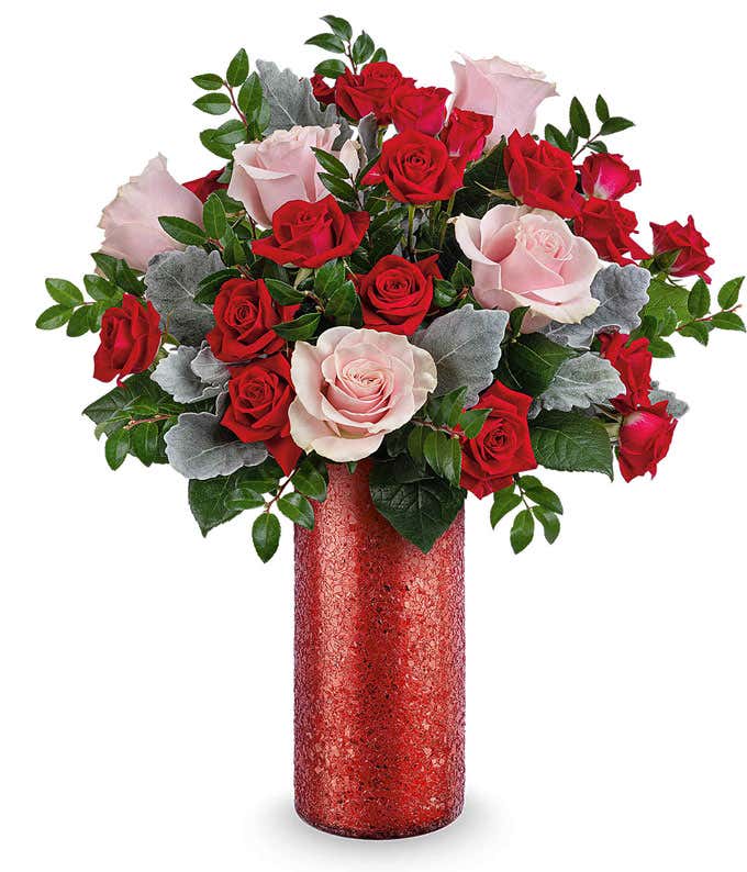 Bouquet of red and blush pink roses with silver dusty miller and lush greenery, arranged in a tall red glitter cylinder vase.