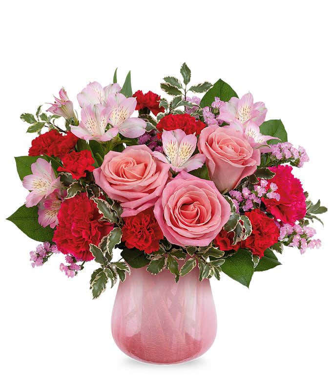 Floral arrangement with soft pink roses, red carnations, pink alstroemeria, delicate filler flowers, and lush greenery arranged in a pink glass vase.