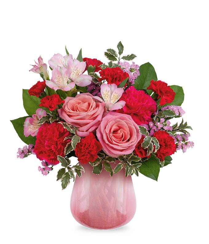 Floral arrangement with soft pink roses, red carnations, pink alstroemeria, delicate filler flowers, and lush greenery arranged in a pink glass vase.