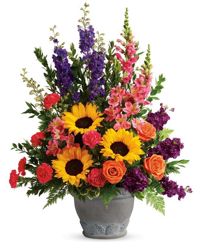 A sympathy arrangement featuring Orange Roses, Pink Alstroemeria, Hot Pink Carnations, Yellow Sunflowers, Purple Larkspur, Hot Pink Snapdragons, Purple Stock, and Floral Greenery in a Grey Ceramic Pot, offering comfort and solace during times of sorrow.