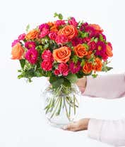Alternative image 4 for Floral arrangement featuring light orange roses, orange spray roses, hot pink carnations, and matsumoto asters in a clear glass vase.