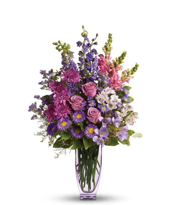 A beautiful lavender and pink arrangement in a lavender vase featuring roses, alstroemeria, snapdragons, larkspur, freesia, matsumoto asters, monte cassino asters, cushion spray mums, statice, and limonium. This elegant display offers a harmonious and soo