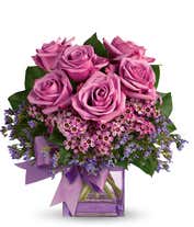 A striking arrangement featuring purple roses, seasonal greens, elegantly presented in a square glass vase adorned with a purple ribbon. This beautiful combination offers a luxurious and sophisticated display, perfect for any special occasion.