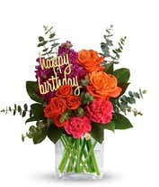 Floral arrangement featuring orange roses, orange spray roses, fuchsia stock, pink carnations, spiral eucalyptus, and lemon leaf with a 'Happy Birthday' pick in a clear cube vase.
