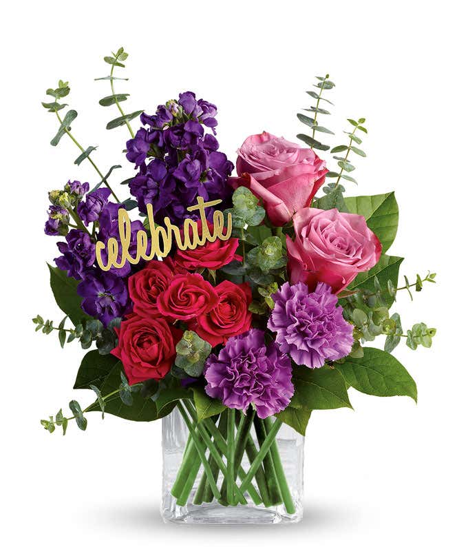 A floral arrangement featuring lavender roses, hot pink spray roses, purple stock, lavender carnations, spiral eucalyptus, and lemon leaf in a glass cube, with a 'Celebrate' pick.