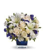 Floral arrangement featuring crème roses, crème spray roses, white Asiatic lilies, white miniature carnations, and purple statice in a blue cube vase.