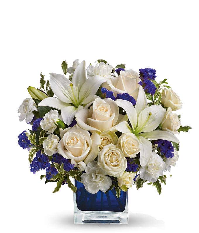 Floral arrangement featuring crme roses, crme spray roses, white Asiatic lilies, white miniature carnations, and purple statice in a blue cube vase.