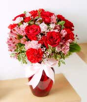 Alternative image 1 for A striking arrangement featuring red roses, pink alstroemeria, red carnations, and pink waxflower, elegantly presented in a red vase with a ribbon. This bold mix offers a stunning and romantic display.