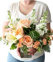 Alternative image 4 for Floral arrangement featuring peach roses, white Asiatic lilies, peach mini carnations, white stock, pitta negra, dusty miller, and a green echeveria succulent in a glass cube vase.