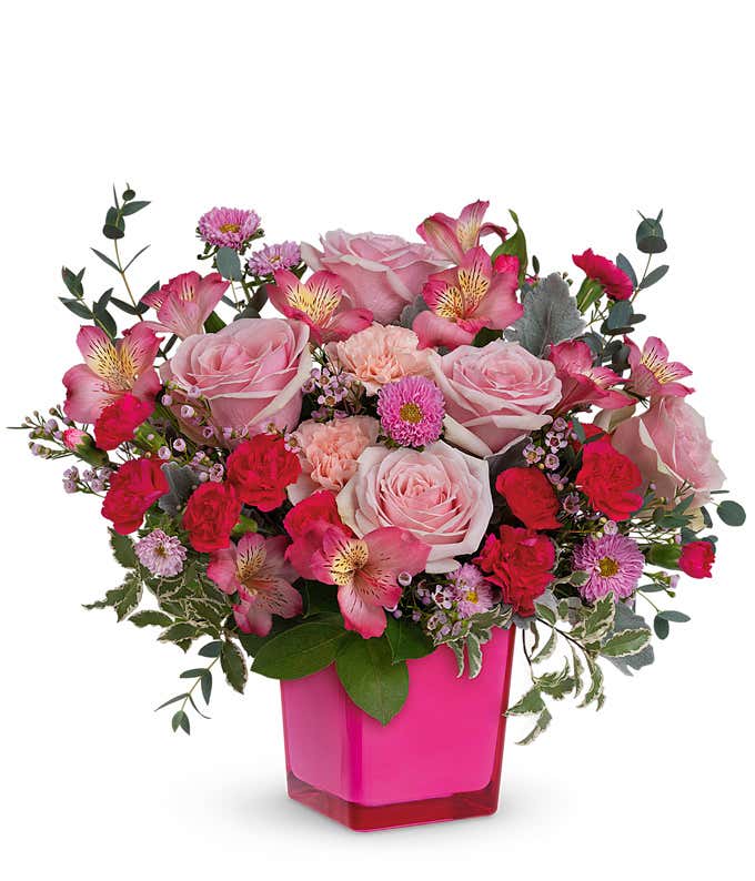Short arrangement of flowers in various shades of pink, placed in a pink cube vase