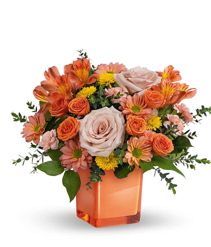 Light orange and coral roses, peach alstroemeria, peach carnations, Yellow and bronze chrysanthemums, pravifolia eucalyptus, lemon leaf in a glass coral cube vase against a white background