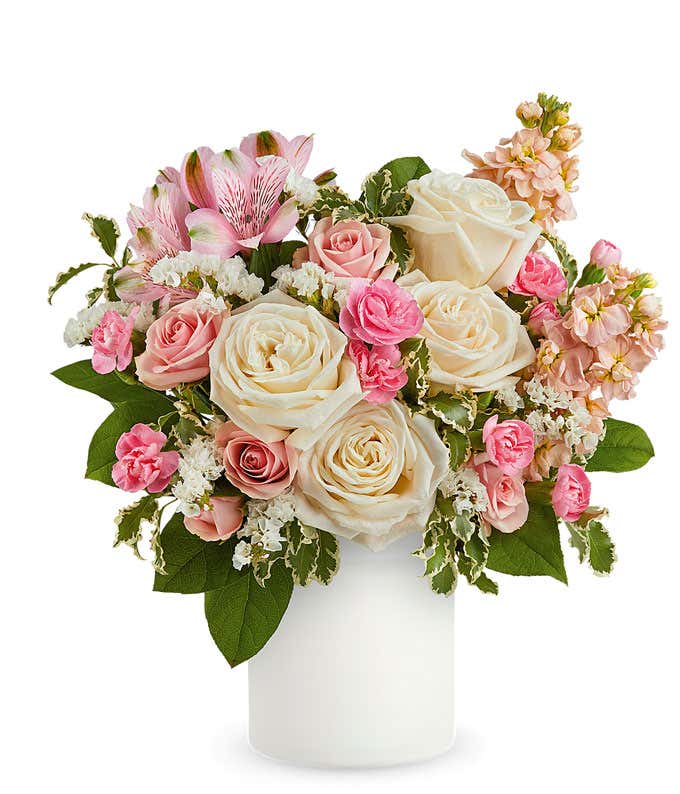 Delicate floral arrangement with ivory roses, light pink spray roses, pink alstroemeria, and pale peach stock in a sleek white vase, perfect for expressing love and care.