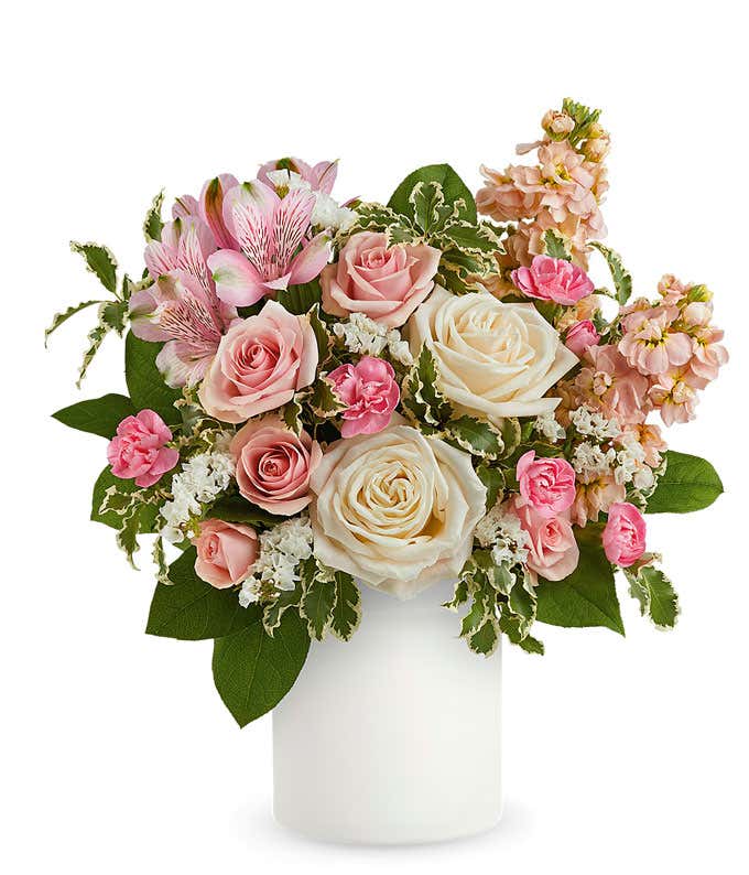 Delicate floral arrangement with ivory roses, light pink spray roses, pink alstroemeria, and pale peach stock in a sleek white vase, perfect for expressing love and care.