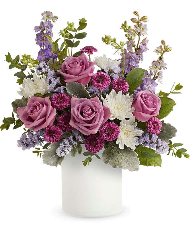 Image of a Mother's Day arrangement featuring Lavender Roses, Purple Button Chrysanthemums, Lavender Larkspur, Lavender Sinuata Statice, floral greens, all beautifully presented in a keepsake White Vase.