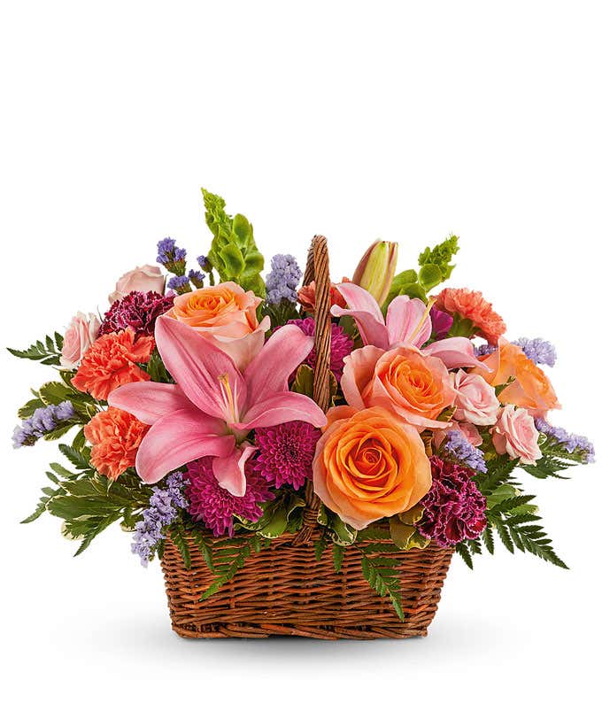 Image of a Mother's Day arrangement featuring Orange Roses, Light Pink Spray Roses, Pink Asiatic Lilies, Burgundy Carnations, Orange Carnations, Purple Cushion Spray Carnations, Blue Sinuata Statice, floral greenery, all beautifully presented in a wicker 