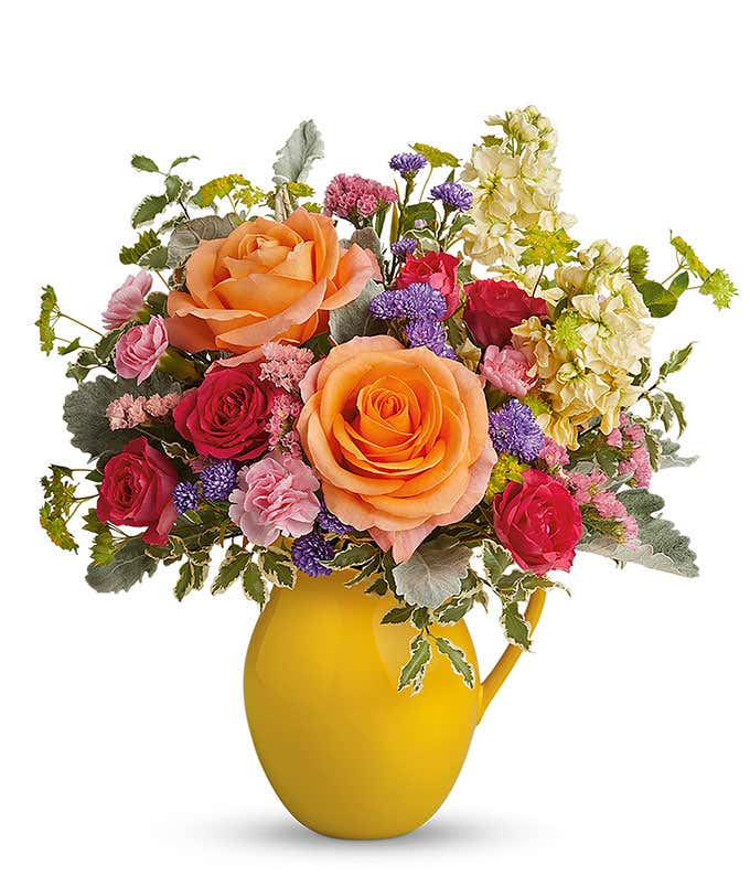A cheerful bouquet of peach roses, hot pink spray roses, miniature pink carnations, yellow stock, and purple monte cassino, arranged in a keepsake yellow pitcher. A perfect gift for any occasion.