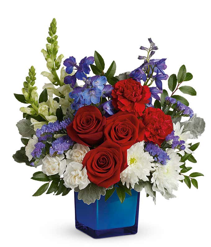 Image of Memorial Day arrangement featuring red roses, red carnations, white miniature carnations, blue delphinium, white snapdragons, white cushion chrysanthemums, and lush floral greenery in a blue glass cube vase.