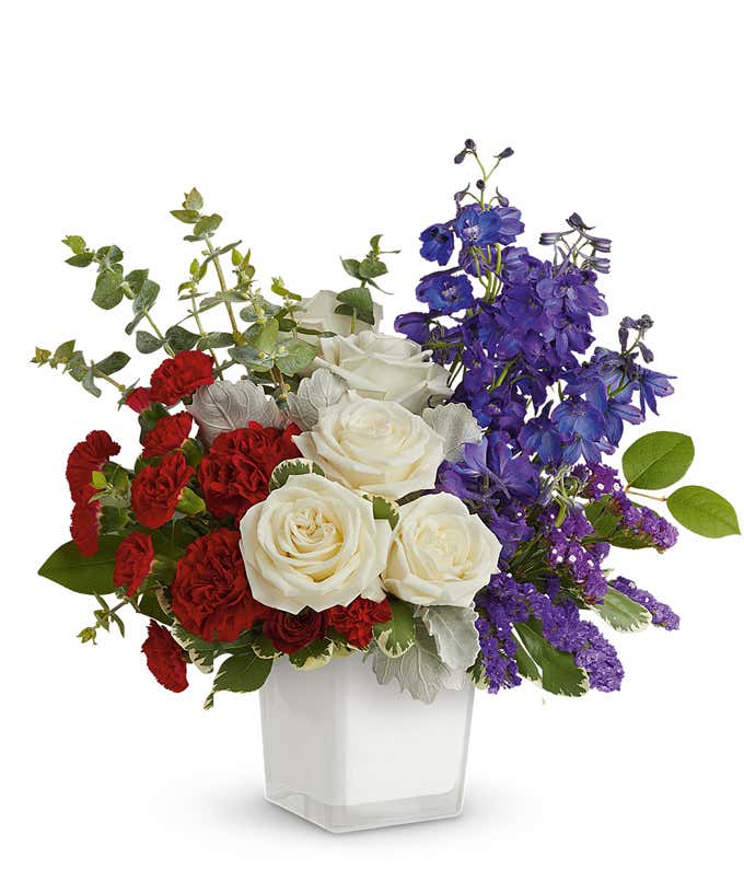 Image of Memorial Day arrangement featuring white roses, red roses, red miniature carnations, blue delphinium, blue sinuata statice, and lush floral greens in a white cube vase.