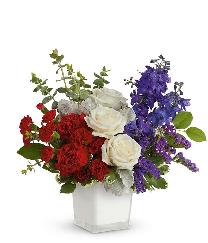 Image of Memorial Day arrangement featuring white roses, red roses, red miniature carnations, blue delphinium, blue sinuata statice, and lush floral greens in a white cube vase.