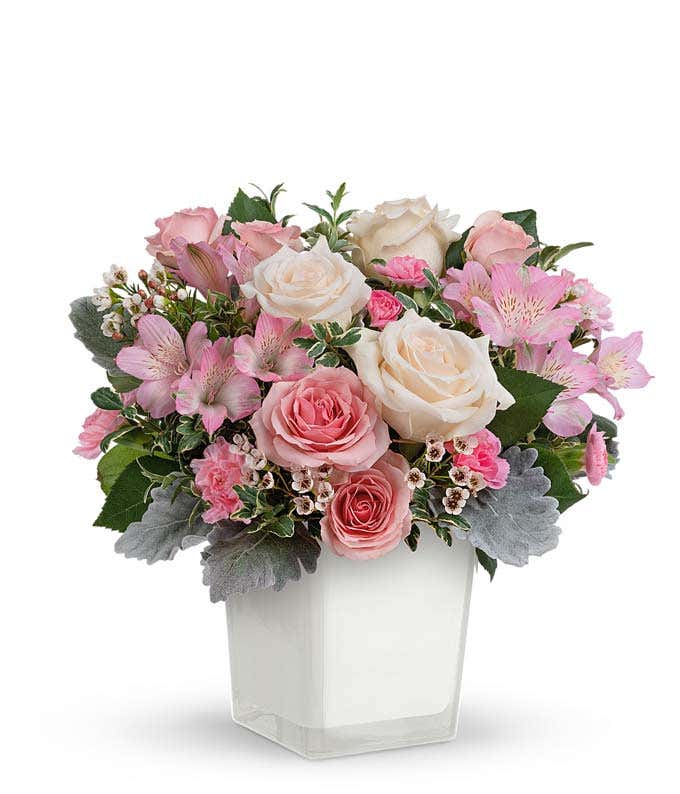 A floral arrangement featuring crème roses, pink spray roses, pink alstroemeria, pink miniature carnations, white waxflower, and greenery in a white vase.