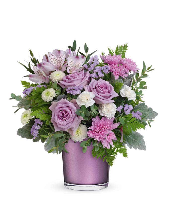 A floral arrangement of lavender roses, alstroemeria, miniature white carnations, white button spray chrysanthemums, lavender cushion spray chrysanthemums, and greenery in a purple cylinder container.