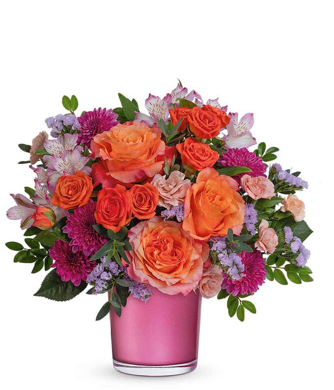 Vibrant floral arrangement with orange roses, orange spray roses, purple chrysanthemums, pink alstroemeria, and lavender statice in a shimmering pink vase, perfect gift for loved ones.