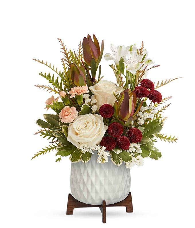 A bouquet of crme roses, white alstroemeria, red leucadendron, peach carnations, and burgundy button spray chrysanthemums in a white vase with a wooden stand.