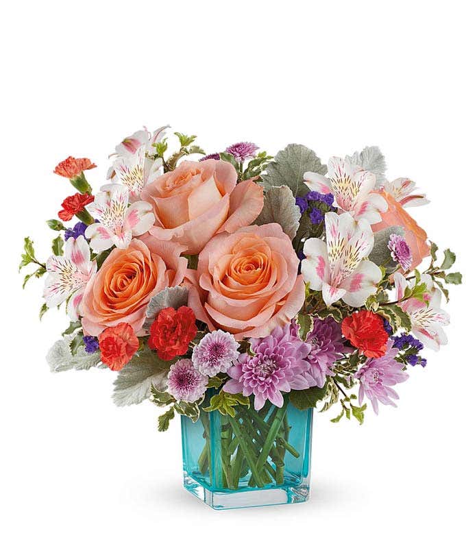 A bouquet featuring peach roses, pink alstroemeria, miniature orange carnations, and lavender cushion spray chrysanthemums in a blue glass vase.