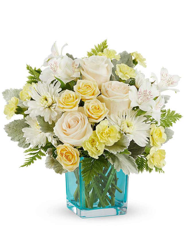 Cheerful floral arrangement with creamy white roses, soft yellow spray roses, white alstroemeria, white mums, and yellow carnations in a vibrant blue glass vase, perfect gift for loved ones.