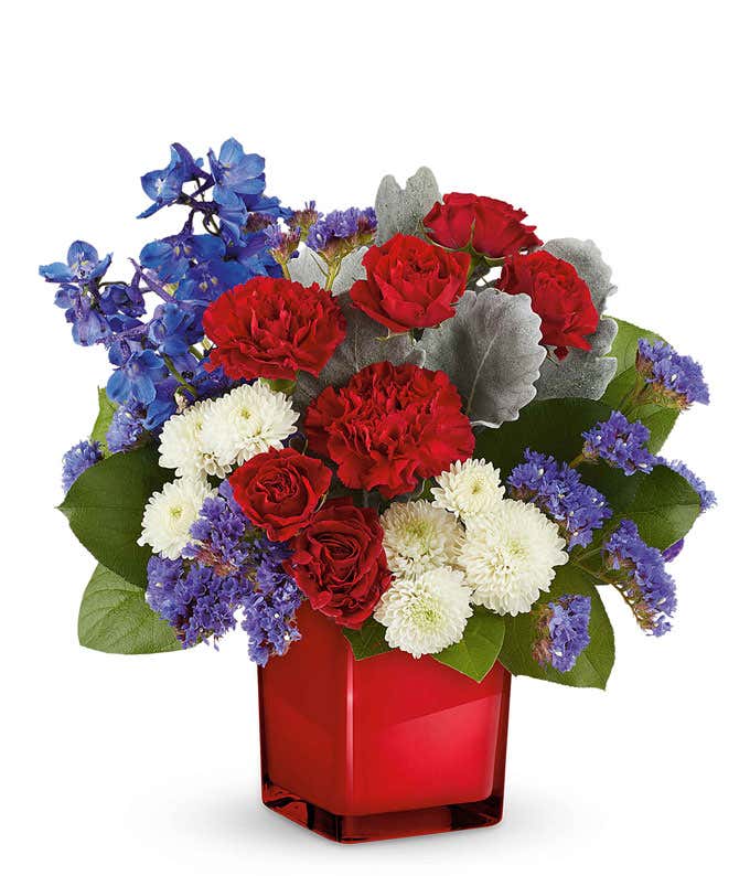 Patriotic floral arrangement with red roses, red carnations, blue delphiniums, blue statice, and white chrysanthemums in a sleek red glass vase, perfect gift for celebrating national pride.