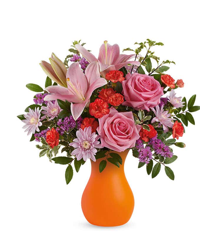 Vibrant floral arrangement featuring pink roses, pink Asiatic lilies, orange spray roses, red miniature carnations, lavender cushion spray chrysanthemums, purple statice, and lush greenery in a bold orange vase.