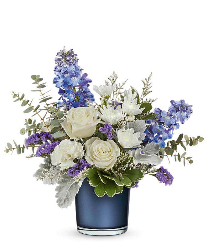 Elegant floral arrangement featuring white roses, white poms, deep blue delphinium, purple statice, dusty miller, silver dollar eucalyptus, and assorted greenery in a sleek navy blue glass vase.