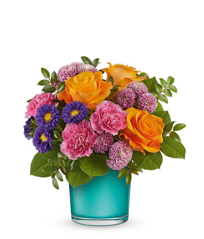 Vibrant floral arrangement featuring orange roses, pink carnations, purple asters, lavender poms, and lush greenery in a turquoise glass container.