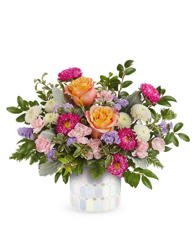 Elegant floral arrangement featuring peach and orange roses, pink spray roses, hot pink Matsumoto asters, white poms, lavender statice, and assorted greenery in a pastel hexagon-patterned ceramic container.